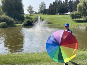Mona mit dem Regenbogenschirm auf einem Golfplatz von einem Wasser mit Fontäne. 