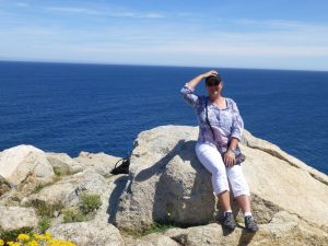 Mona auf einem Felsen auf Korsika sitzend. Mit den Händen auf der Kappe, damit sie nicht vom Wind verweht wird. Im Hintergrund das Meer. 